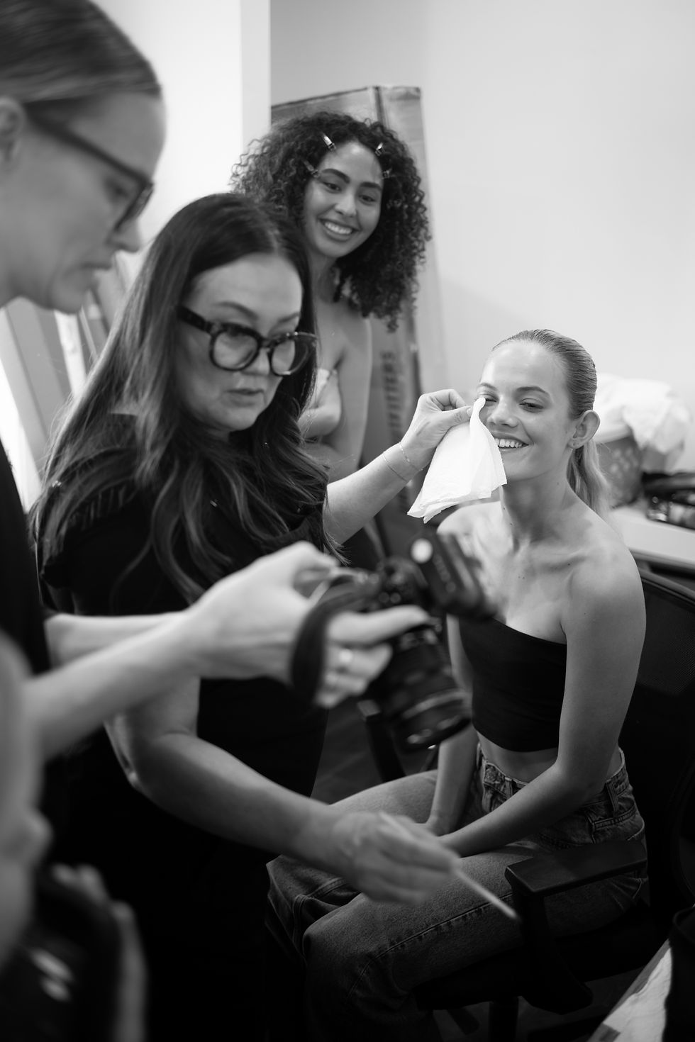 Woman in black top smiles as another person wipes her face. Two others observe, one holding a camera. Indoor setting, relaxed mood.