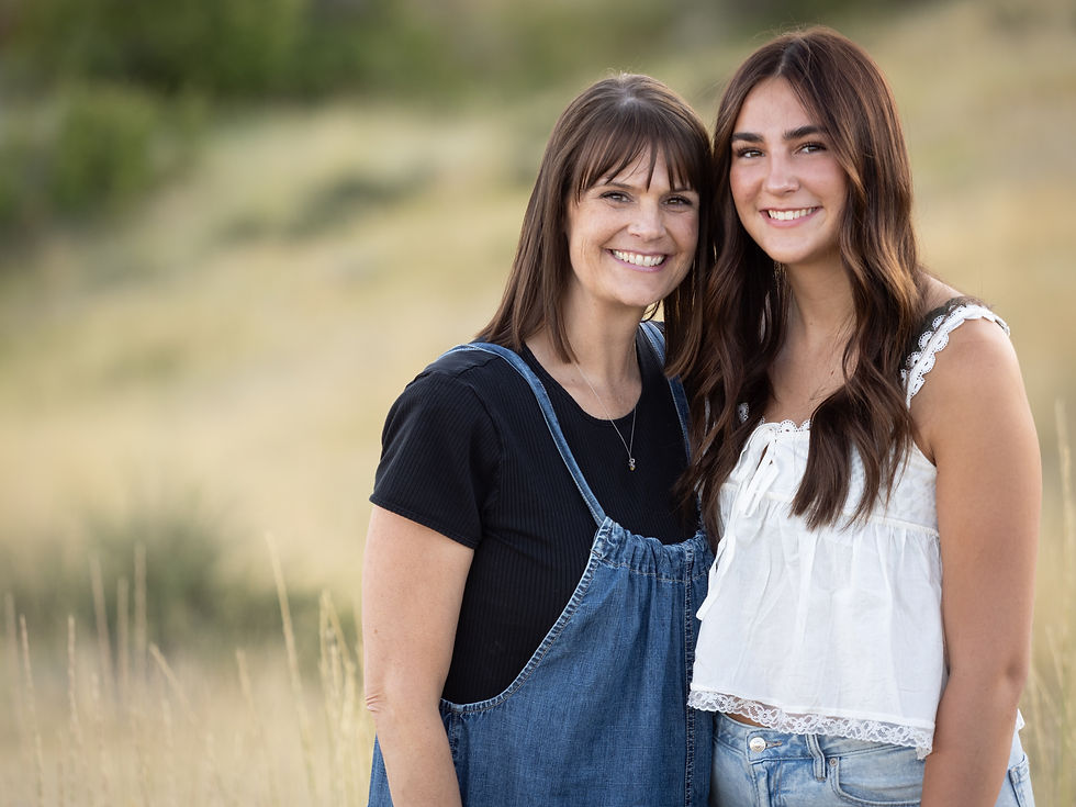 Two women smiling in a grassy field. One in a black top with denim overalls, the other in a white lace top. Bright, cheerful mood.