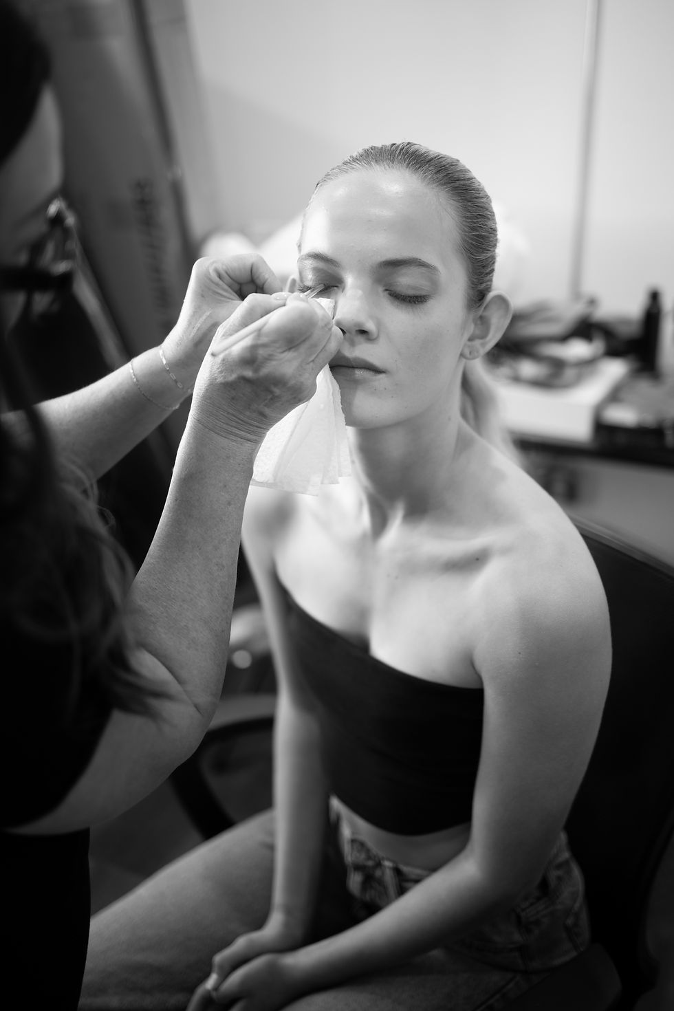 Makeup artist applies eye makeup to a seated woman in a black top. The setting is a bright room, and the scene is calm and focused.