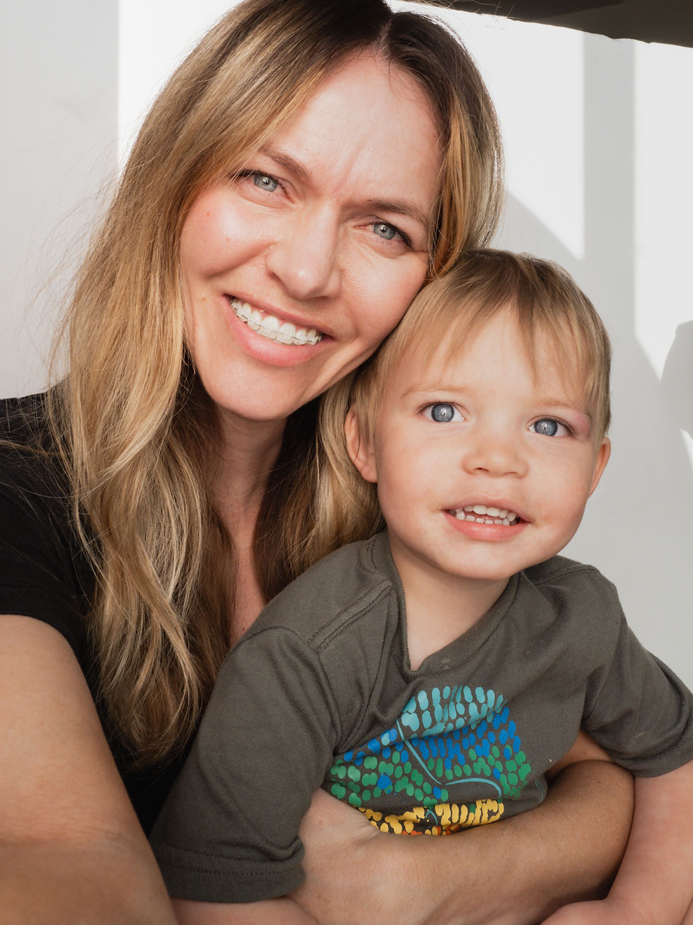 Smiling woman and child in close embrace, with natural light background. Child wears a gray shirt with colorful print. Happy and warm mood.