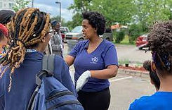 a light skinned black woman ( yordanose) wearing a blueshirt, gardening gloves talking to a group of youth about instructions for planting