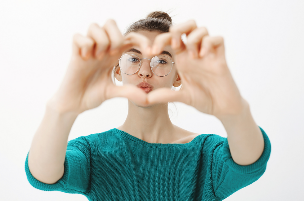 A woman with her hair in a bun, wearing large, round metal-rimmed glasses and a teal sweater, holding her hands up to form a heart shape with her fingers. She is looking through the heart.