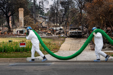 CCC Corpsmembers carry a large compost sock past burned homes.
