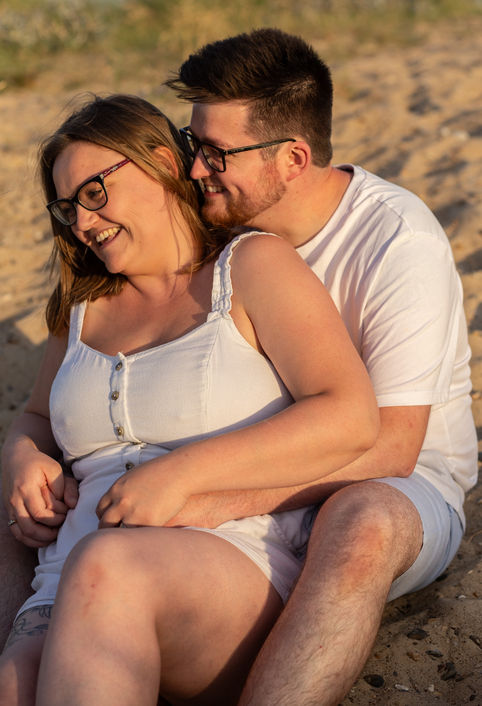 A happy couple sits together on the beach, smiling, wearing glasses.