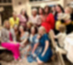 A group of smiling women and children pose in colorful outfits in a room with patterned carpet and large windows, creating a joyful atmosphere.