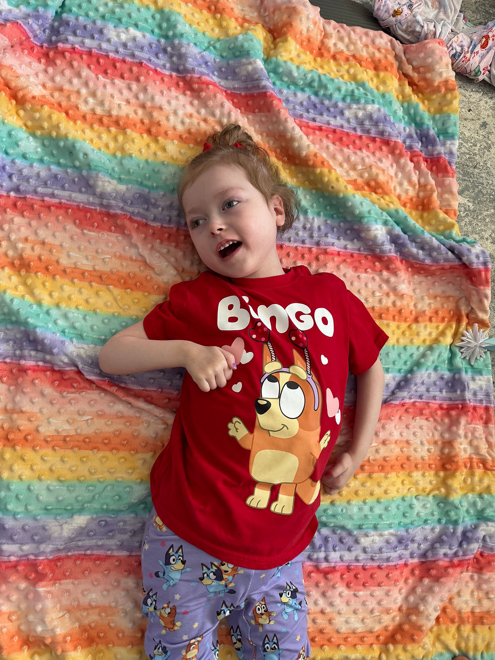 Charlotte lying on a colorful rainbow blanket, wearing a red "Bingo" shirt and colorful pants, smiling with joy.