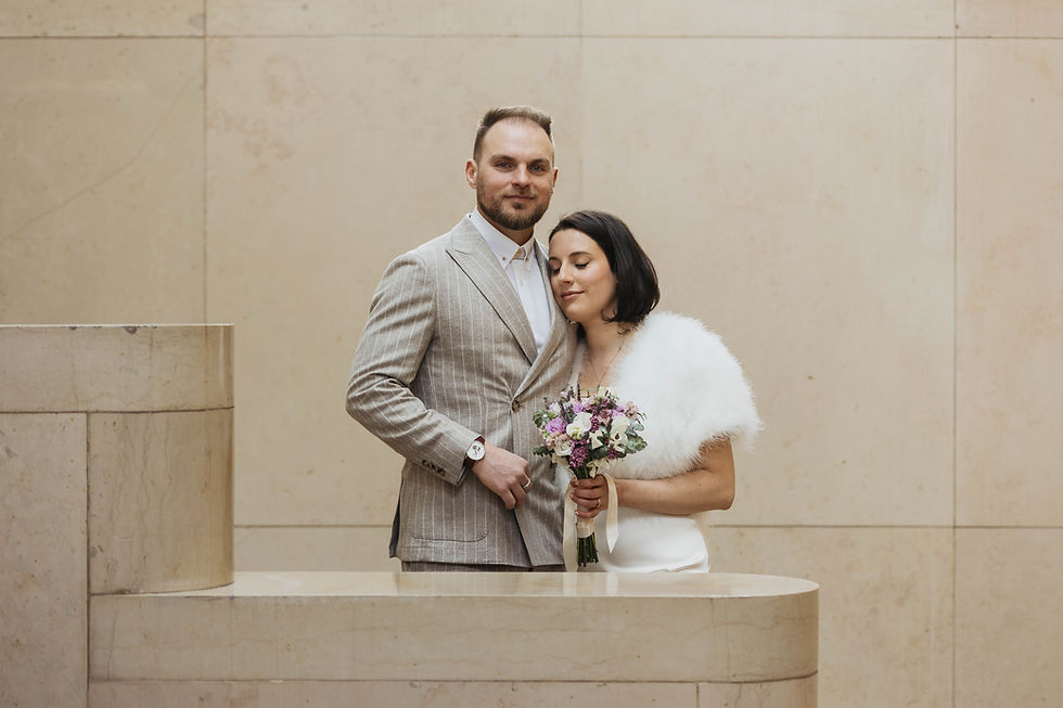Un couple en tenue élégante se tient près d'un mur beige. La femme tient un bouquet fleuri, exprimant la sérénité.