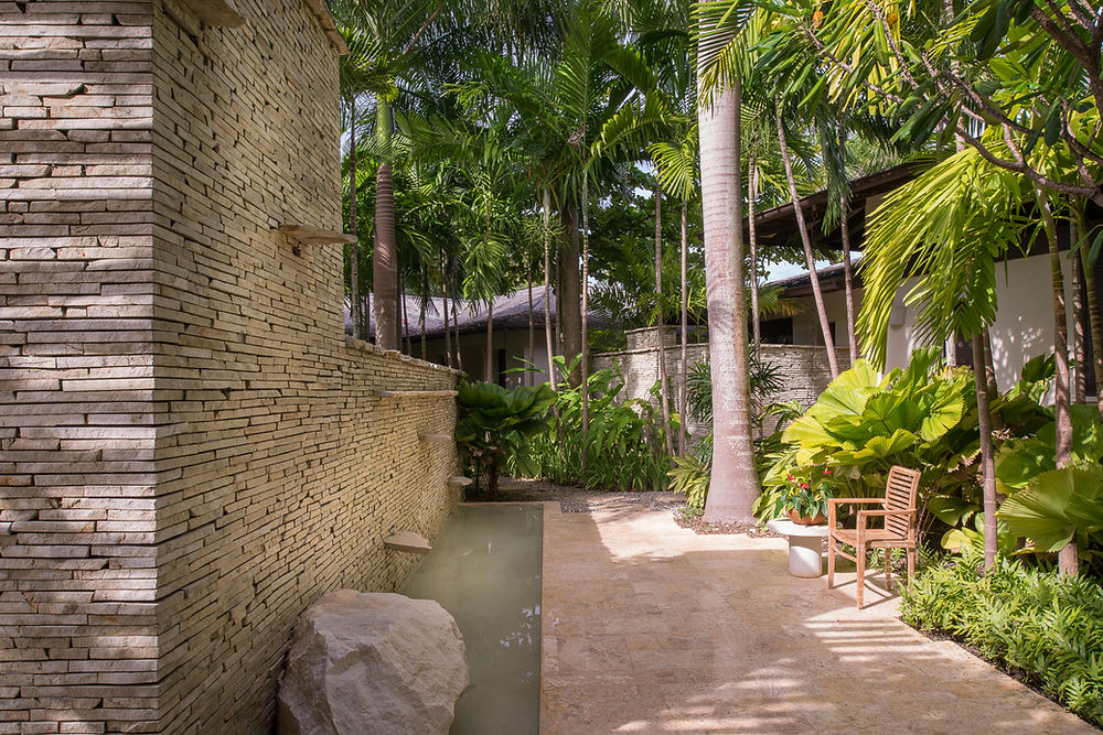 Tropical garden path, stone wall, water feature, and chair.