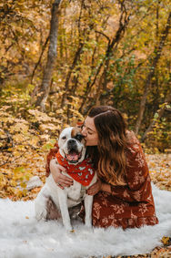 A woman kisses her dog's cheek. They're on a white blanket in a fall forest.