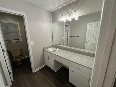 Bathroom with gray walls, large mirror, granite countertop, and vanity lights. Toilet and closed blinds visible in the adjacent room.