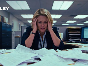 A woman looks stressed, holding her head in an office with piles of papers on the desk. Fluorescent lights, a computer, and "Foley" logo visible.