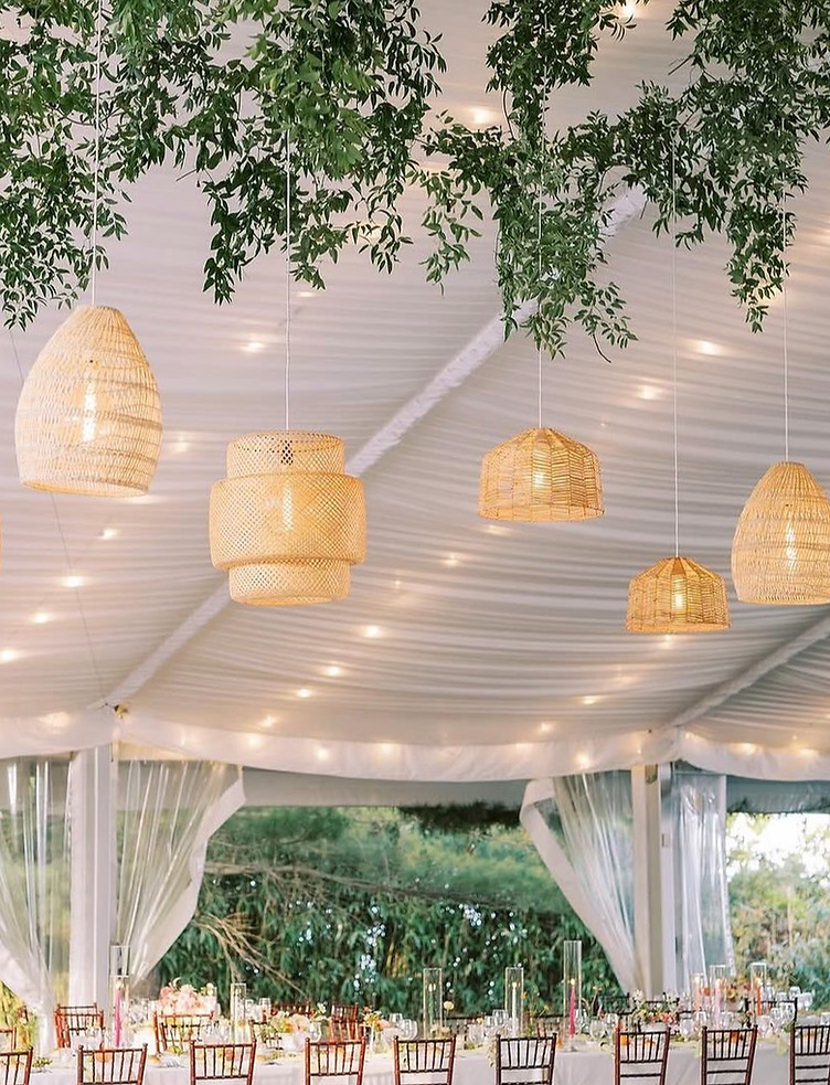 Warm string lights and woven pendant lights illuminate an elegant outdoor tent with flowing white draping and greenery.