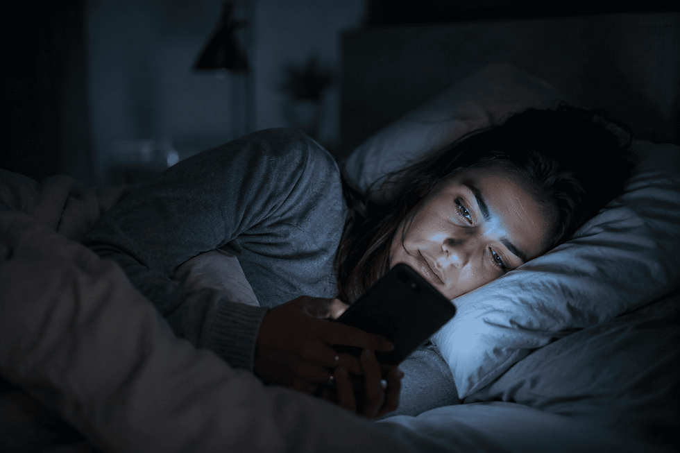 Mulher adulta deitada na cama durante a noite, iluminada pela luz azul do telemóvel, num quarto escuro e minimalista. Ambiente silencioso e intimista que transmite o impacto do uso de ecrãs no sono e no descanso noturno.