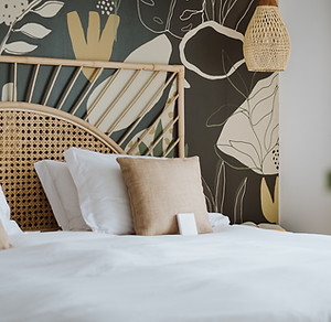 Bedroom with graphic floral wallpaper in beige and ochre tones on a dark background, combined with a rattan headboard and neutral bedding.