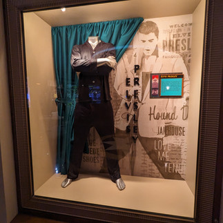 A display case showcases a black outfit worn by Elvis Presley, with the shirt buttoned up and the arms crossed. Behind the outfit, a turquoise curtain complements the background, which features sepia-toned wall art with phrases related to Elvis, including "Jailhouse Rock" and a photo of him. Informative panels with text provide context about the displayed outfit.
