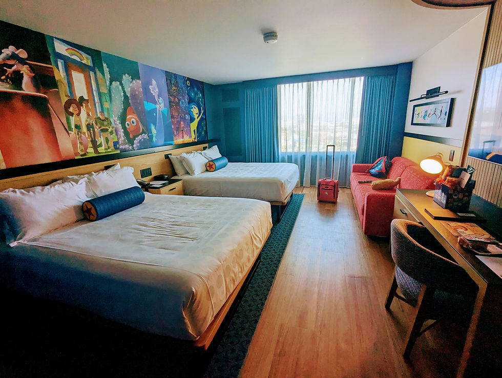 A brightly lit hotel room at Disneyland's Pixar Place Hotel featuring two queen-sized beds with white linens and blue cylindrical pillows. Behind the beds is a wall mural depicting scenes and characters from Pixar and Disney movies such as "Ratatouille," "Toy Story," "Finding Nemo," "Coco" and "Inside Out ". Light blue curtains frame a window overlooking a city view. To the right, a red sofa with throw pillows is positioned near a desk with a lamp and assorted items. A red suitcase stands near the window. The room’s decor is modern and colorful.