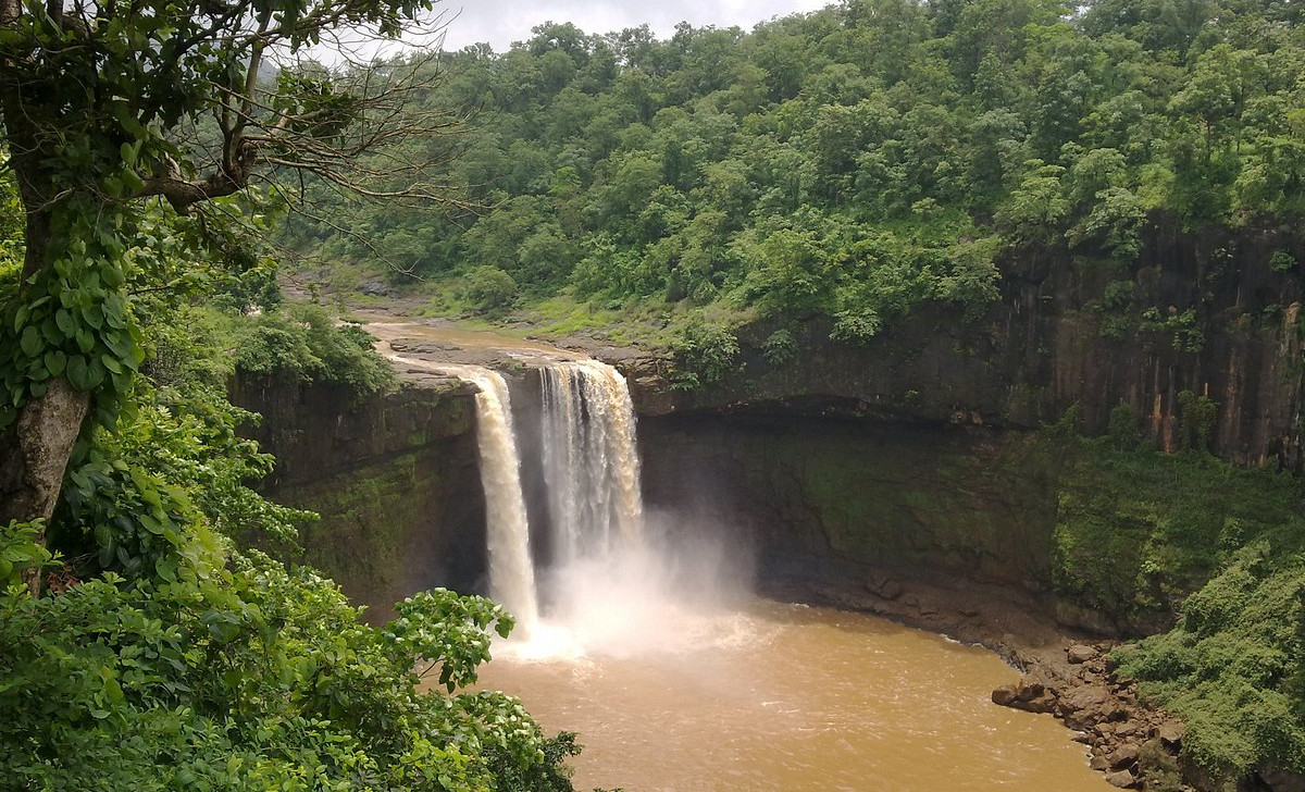 Twin waterfall plunges into muddy pool in a lush green forested gorge under a cloudy sky.