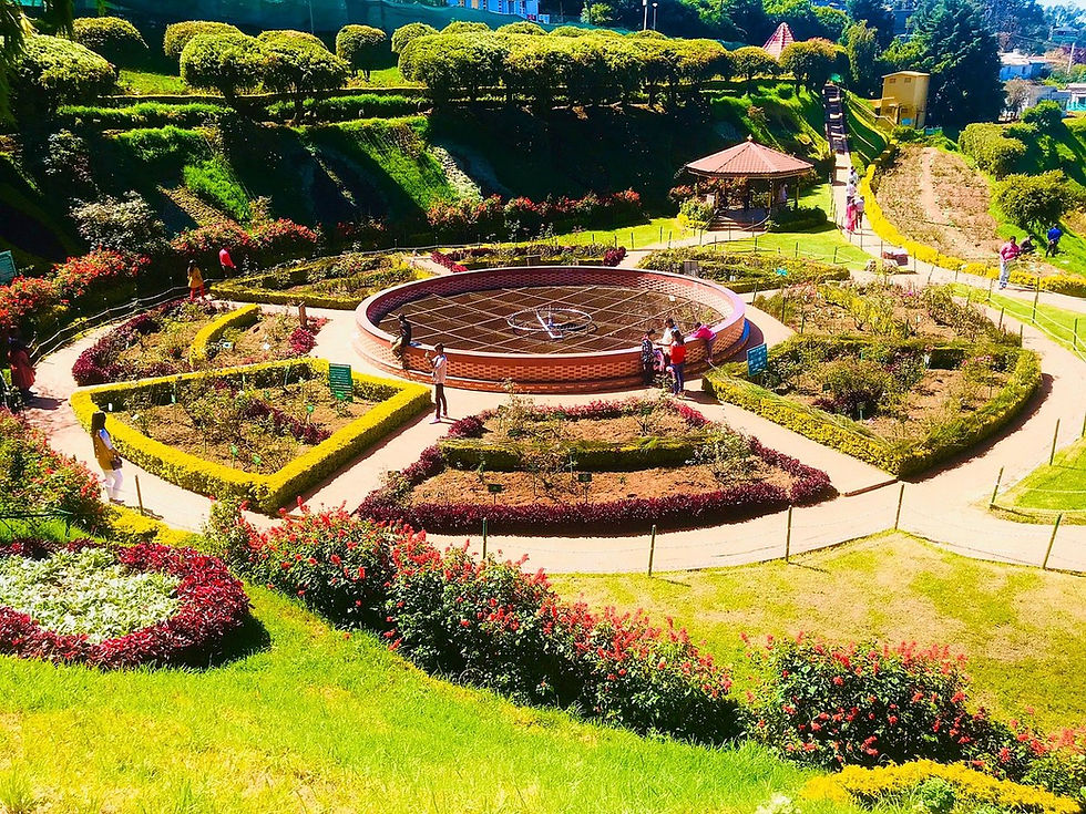 Bright terraced flower garden with winding paths, trimmed shrubs, a gazebo, and visitors walking around a circular central feature