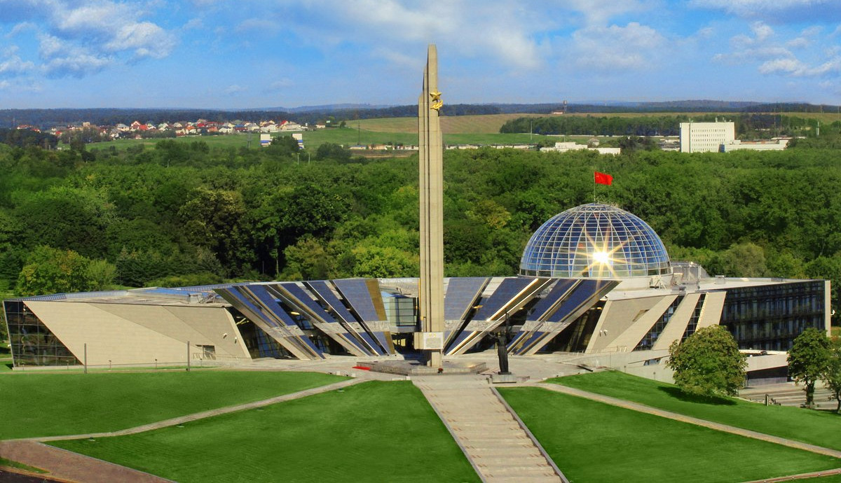 Soviet memorial museum with tall obelisk and glass dome topped by a red flag, set in green park under a bright blue sky.