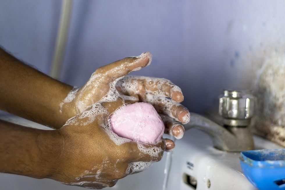 Hands lather with pink soap under a running faucet. Foamy bubbles cover hands. Background has a light blue wall and sink.