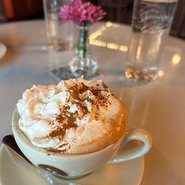 Cup of cappuccino with whipped cream and cinnamon on a table, flanked by two water glasses and a small vase with a pink flower. Cozy setting.