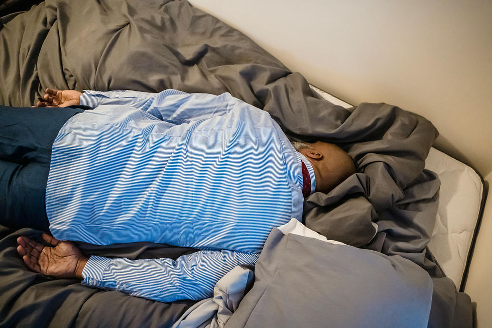 Man in striped shirt lying face down on a bed with gray sheets, appearing to rest. Room has a calm, neutral setting.
