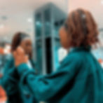A woman in a teal jacket touches her reflection on a glass surface in a modern, softly lit setting. She appears contemplative.