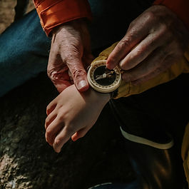 Hands adjust a compass on a child's wrist in a wooded setting. The child wears yellow. The adult wears an orange sleeve, conveying guidance.