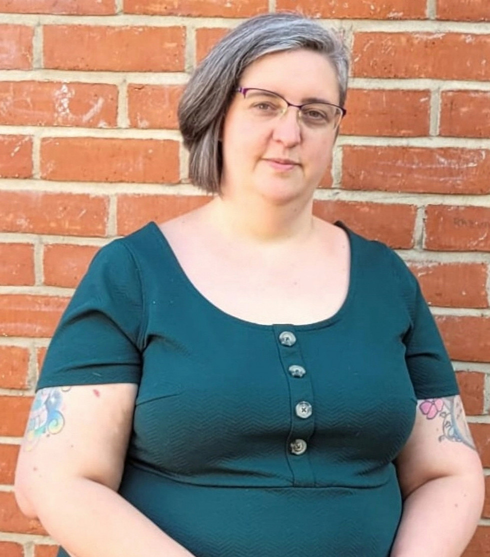 Jenni - a plus-sized white woman, with short brown hair and glasses. She is wearing a dark green dress. She has tattoos on her arms. She is stood in front of a brick wall