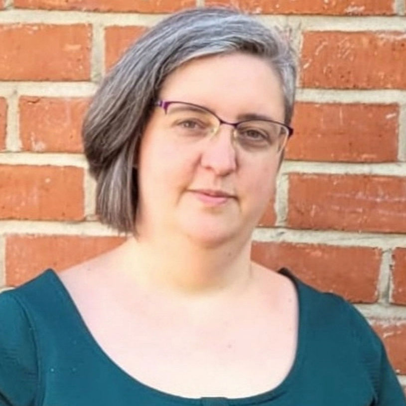 Close up of Jenni - a white woman with short brown hair and glasses. She is in a dark green dress, in front of a brick wall.