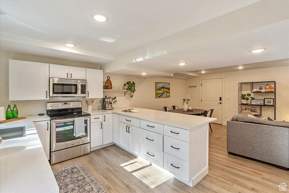 Modern kitchen and living area with white cabinets, stainless steel appliances, and light wood floors. A cozy gray couch and dining table.