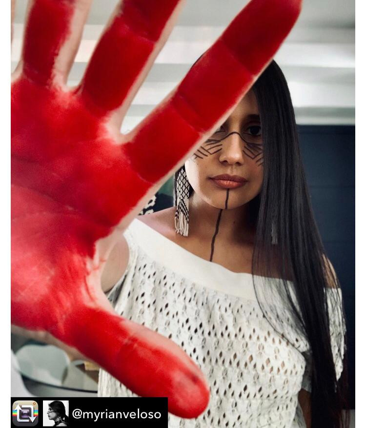 #descricaodeimagem: Dra. Myrian Veloso, ou Krexu, como a chamam na aldeia Rio das Cobras, que significa Lua Crescente, formou-se como médica pela faculdade de medicina da Universidade Estadual do Oeste do Paraná (Unioeste). Ela denuncia o assassinato de lideranças em seu perfil. Na foto, a médica está com o rosto pintado com traços geométricos pretos e a palma da mão direita em destaque, pintada de vermelho bem vivo. Ela tem a pele morena, cabelos pretos compridos e usa franja