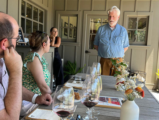 A seated wine tasting in Napa Valley. A winery owner speaks to guests at a wood table outside in a covered area close to a vineyard. Descriptions of the wine served are on a menu in front of each person. A woman leans on a nearby counter, smiling. Flowers complete the bright and cozy experience.