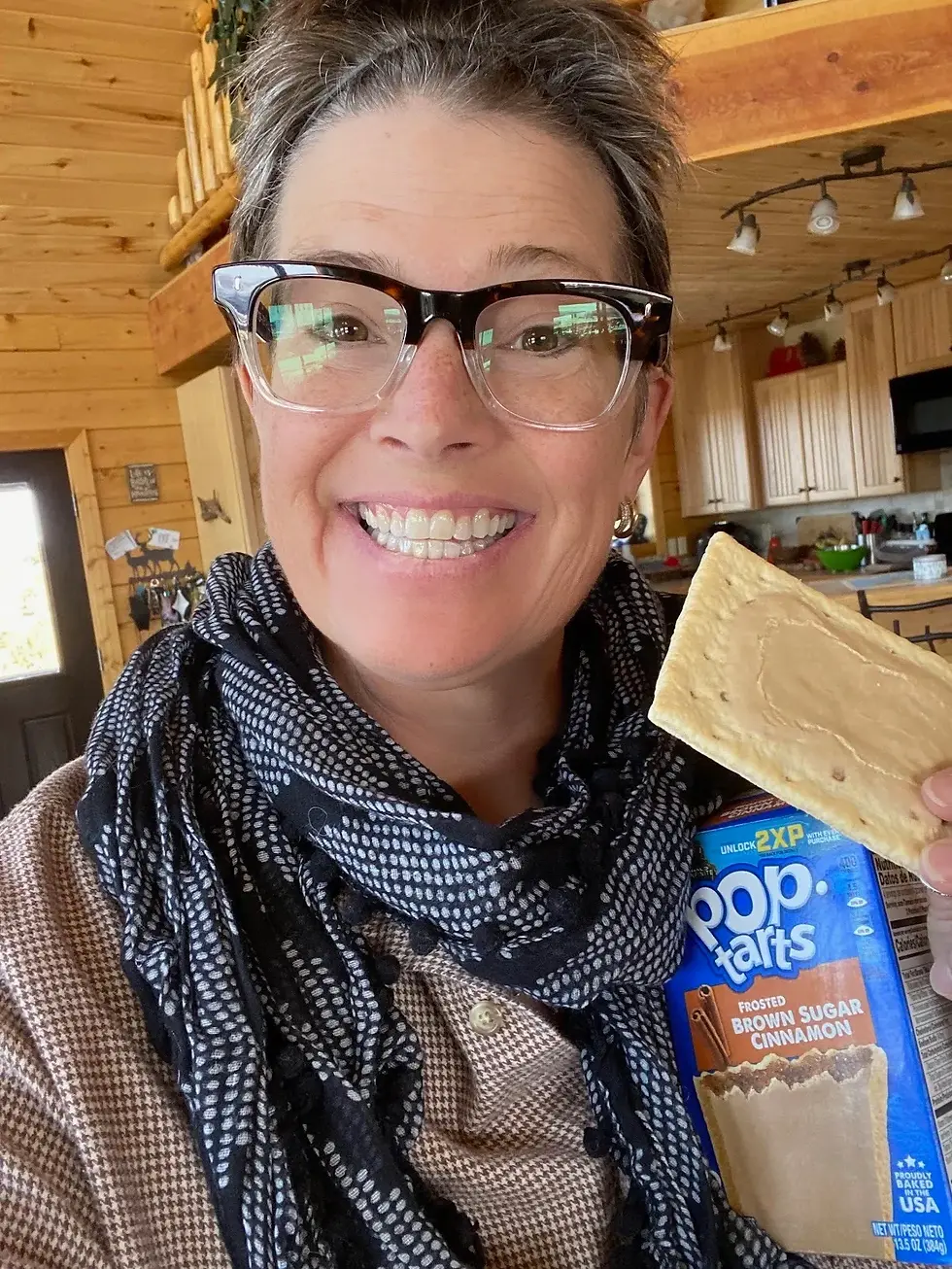 CC is pictured smiling with horn-rimmed glasses, a flannel shirt and black and white polka dotted scarf and holding up a cinnamon brown sugar pop tart and the box.