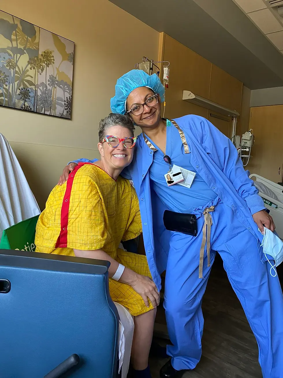 CC sitting in a yellow hospital gown with her female, Indian surgeon in blue scrubs both wearing glasses and hugging in celebration of CC being cancer free