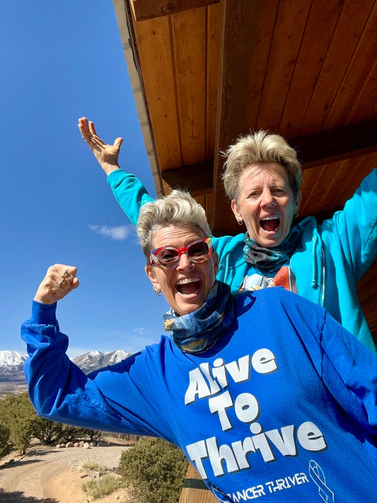 CC and Doc on their deck leaning over the edge: CC with a strong arm selfie and a blue long sleeve shirt that reads, Alive to Thrive. Doc is in an aqua blue shirt with hands in the air celebrating.