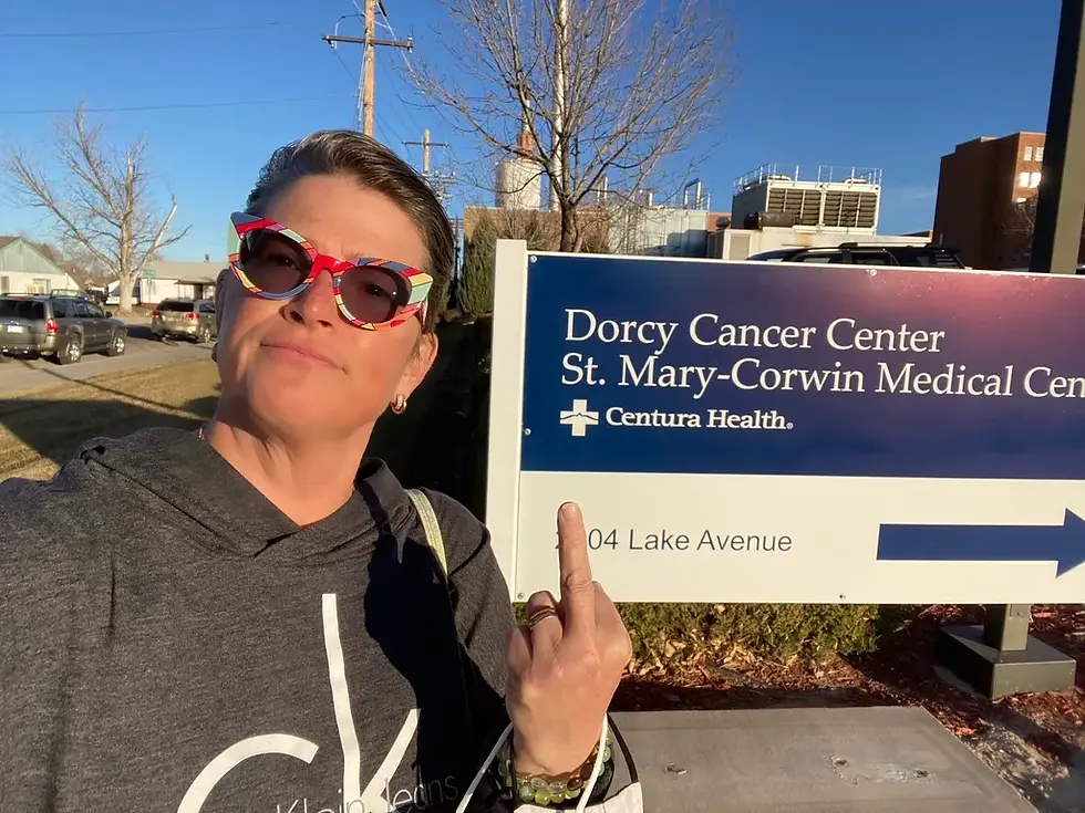 CC stands outside of the Dorcy Cancer Center sign about to go in for her first radiation appointment. She is not thrilled and is holding up her middle finger as if to say fuck cancer. She is dressed in a gray hoodie and red sunglasses.