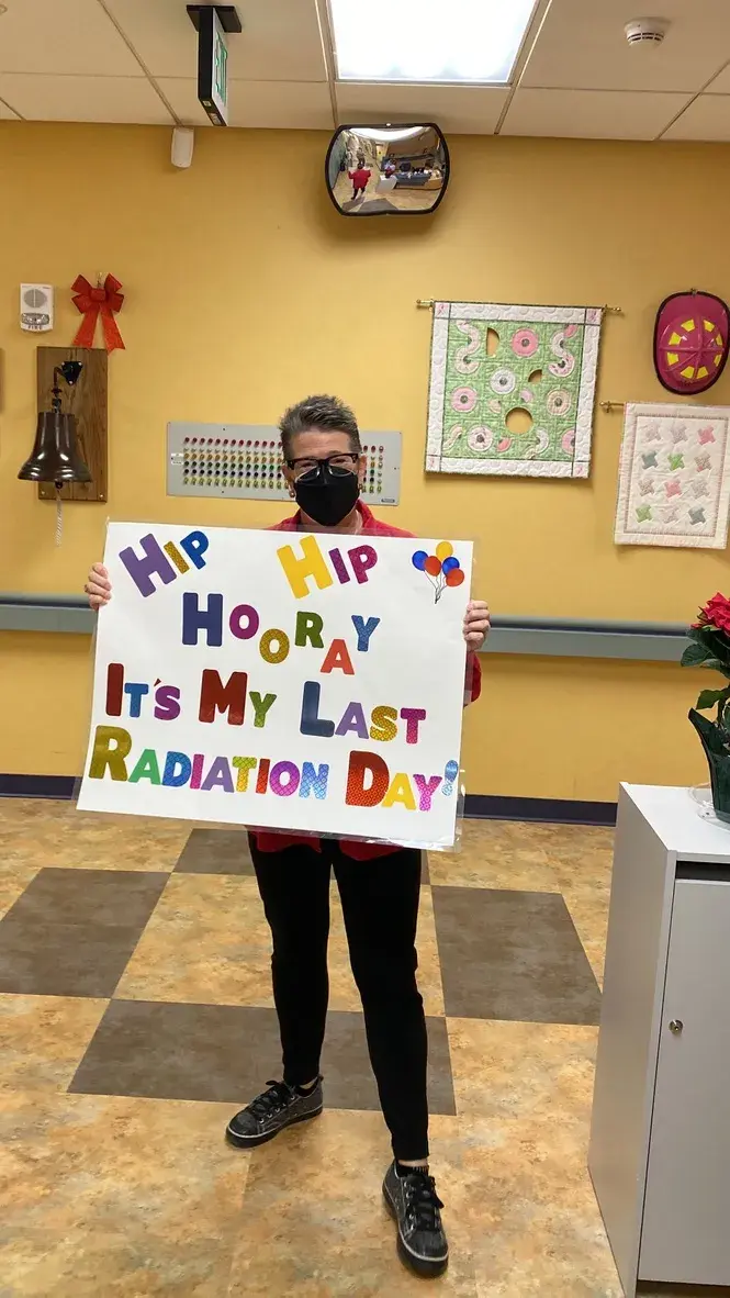 CC holding up a sign that reads, Hip Hip Hooray, It’s my last radiation day! Standing in front of the bell she is about to ring to celebrate the end of radiation!"