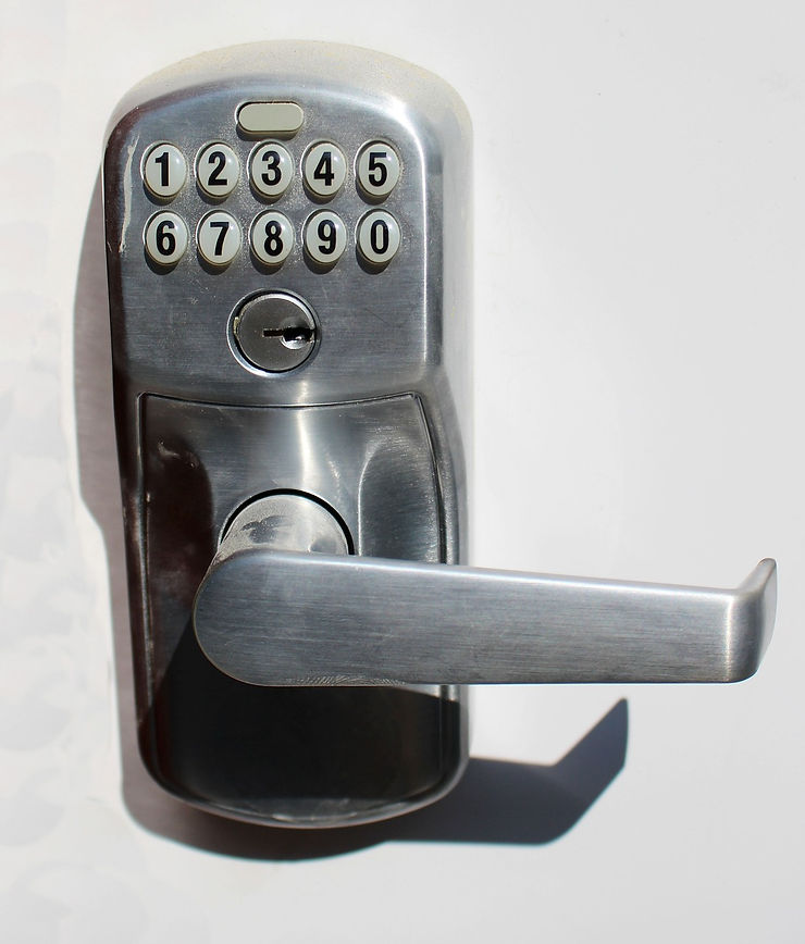 Silver keypad lock with numbers 0-9 on a white door. Sunlight casts a shadow of the handle. A keyhole is visible below the keypad.