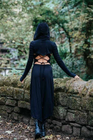 Woman in black dress stands The Fall Cut near stone wall and trees Catana Clothing.