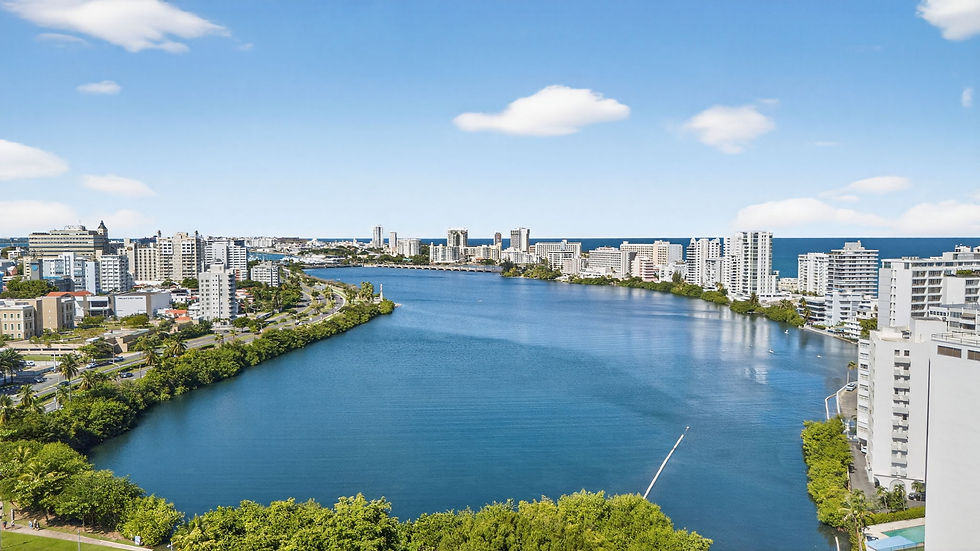 Aerial view of a blue lagoon between white high-rise buildings, with the ocean beyond and a bright sky with scattered clouds.