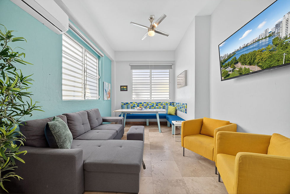 Bright living room with gray sectional, yellow chairs, blue bench, potted plant, and wall art of a waterfront cityscape.