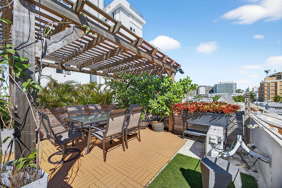 Sunny rooftop patio with pergola, dining table, chairs, and lush potted plants overlooking city buildings under a blue sky