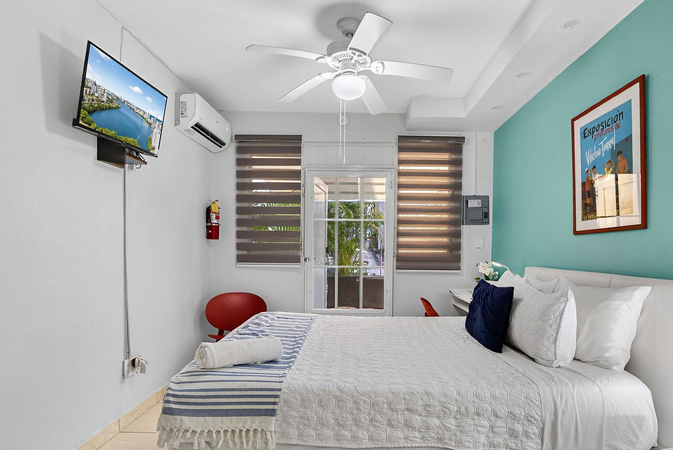 Bright bedroom with white bed, turquoise accent wall, wall TV showing a river cityscape, and a Spanish art poster.