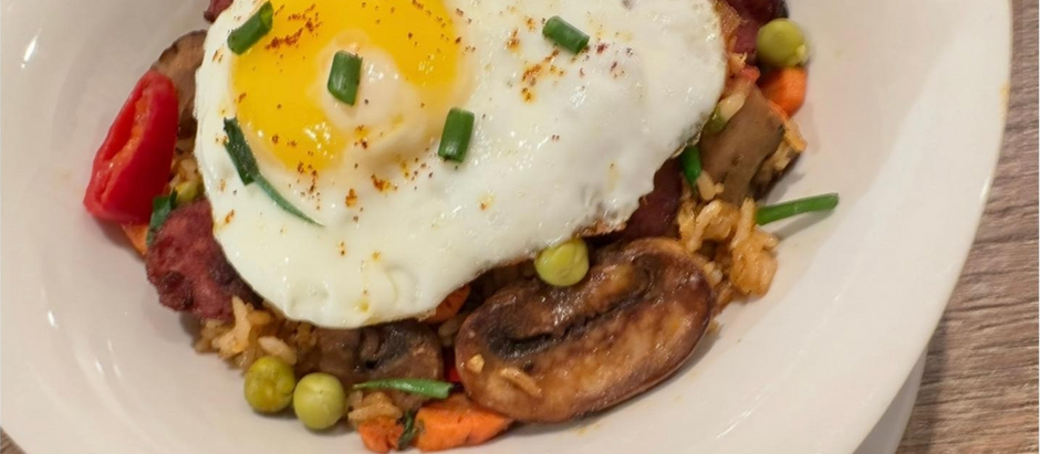 A colorful bowl of seasoned rice mixed with sliced kielbasa, mushrooms, carrots, sweet peas, and red peppers, topped with a sunny-side-up egg and garnished with scallions. A vibrant, Tampa Bay–inspired twist on classic Caribbean rice and eggs.