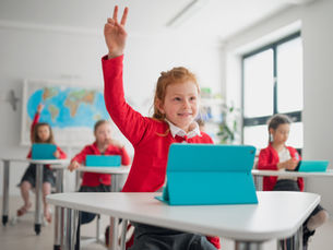 Smiling girl in red raises her hand in a bright classroom, with classmates using blue iPad for education.