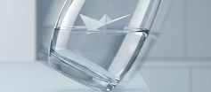 A minimalist editorial illustration shows a white paper boat floating level inside a tilted clear glass that teeters at the edge of a white table. The cool gray setting is calm and elegant, but the unstable composition suggests balance preserved in the midst of imminent risk.