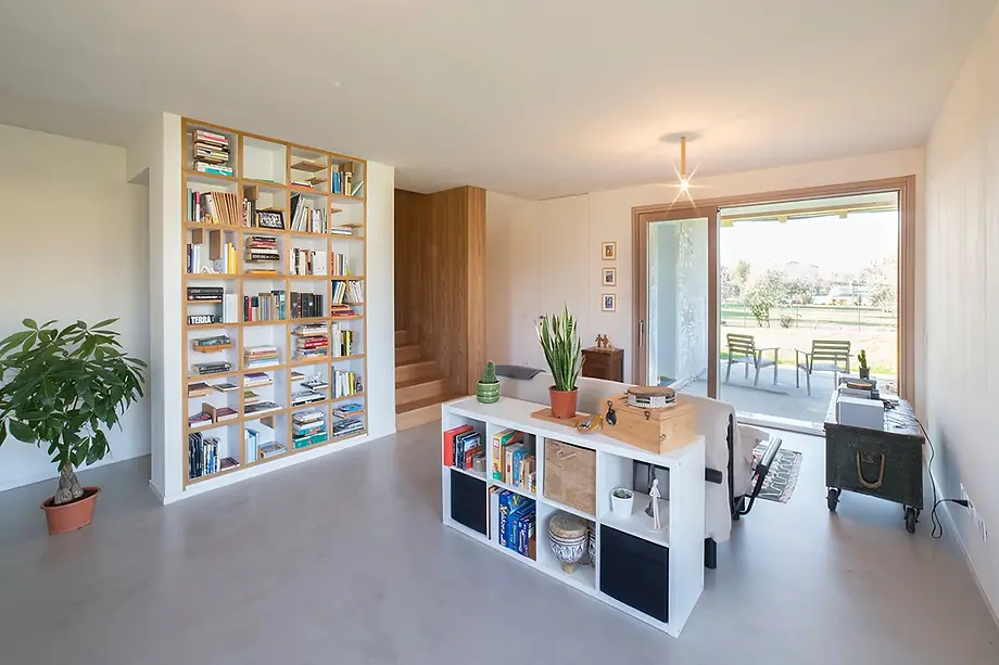 Vista della zona giorno, libreria e mobile a scomparti quadrati e finiture in legno chiaro, cassa panca vintage, divano, ampia vetrata su giardino, vano scale in legno che riveste gradini e pareti.
