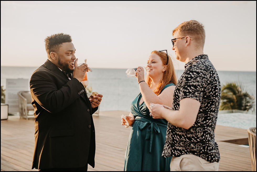 A groom laughs with wedding guests on a resort deck by a Jamaican beach.