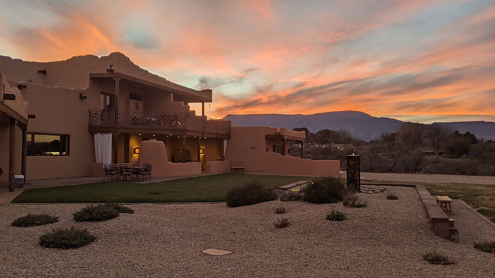 An evening sky of orange and grey clouds silhouettes distant Mingus Mountain. Nearby is a small garden featuring a bubbling stone pillar fountain. To the left is Origins Bed & Breakfast, a Pueblo Revival-style two-story home.